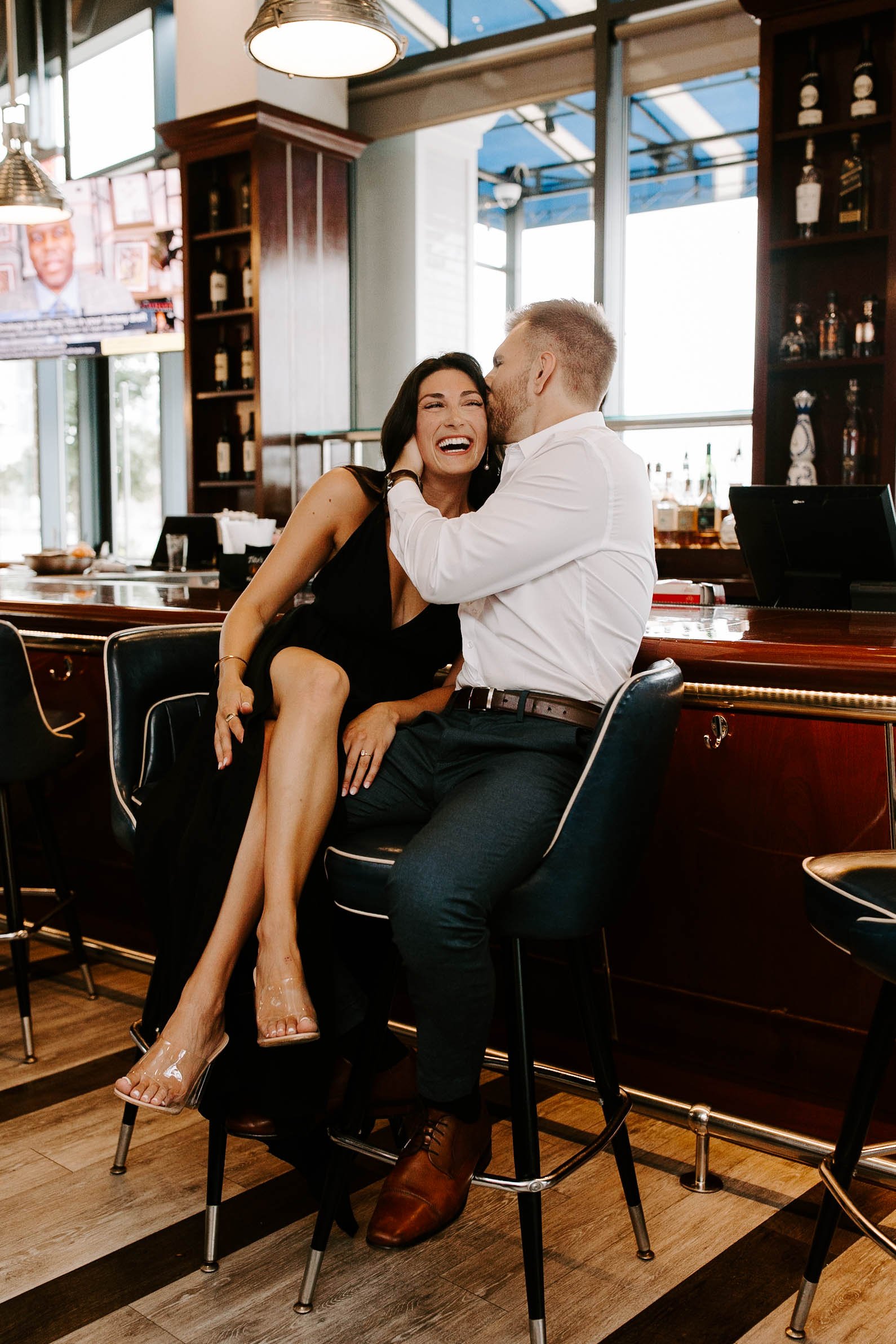  Boyfriend kissing girlfriend’s head while she laughs and smiles while sitting at a restaurant bar  