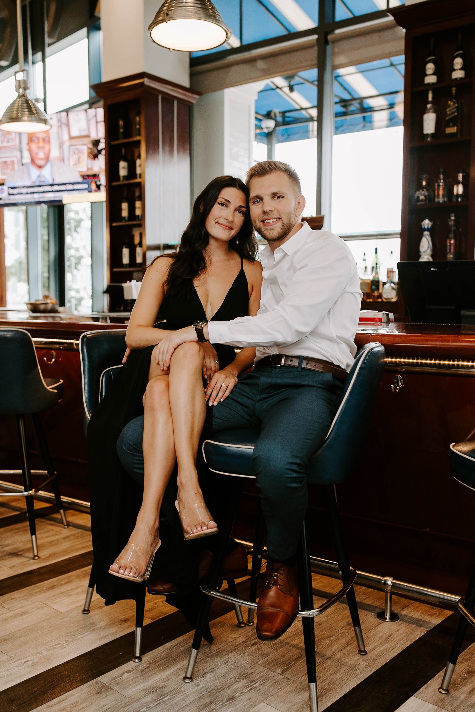  Engaged couple smiling sitting at a restaurant bar, girlfriend wearing a black dress and boyfriend is wearing a white button down and jeans  