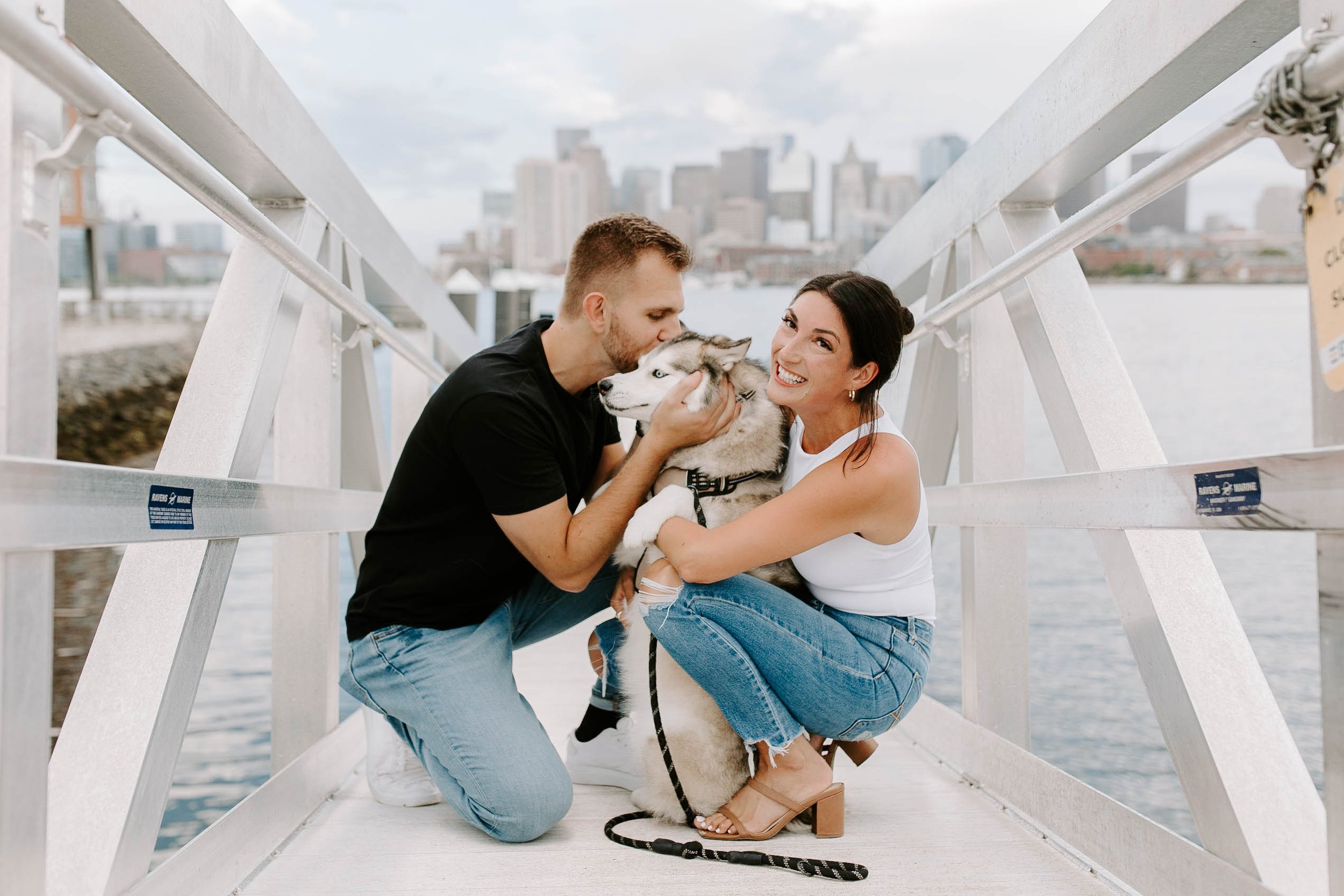 Engaged couple smiling and kissing their husky on the seaport