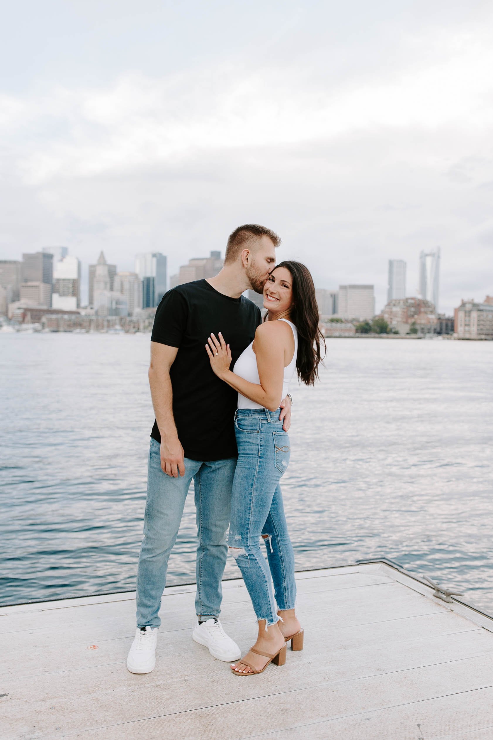  Engaged couple standing on the oceanfront with skyline view in the background, boyfriend kissing fiances forehead 