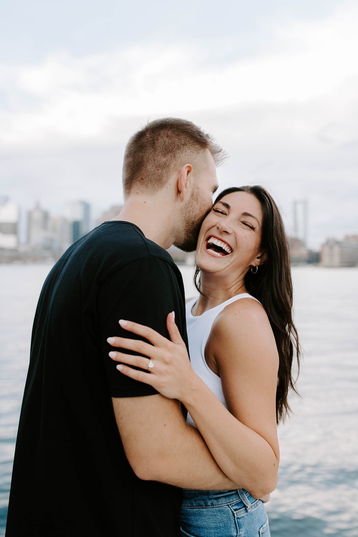  Engaged couple smiling and laughing holding one another at ocean front in front of skyline view  for their boston Seaport engagement session 