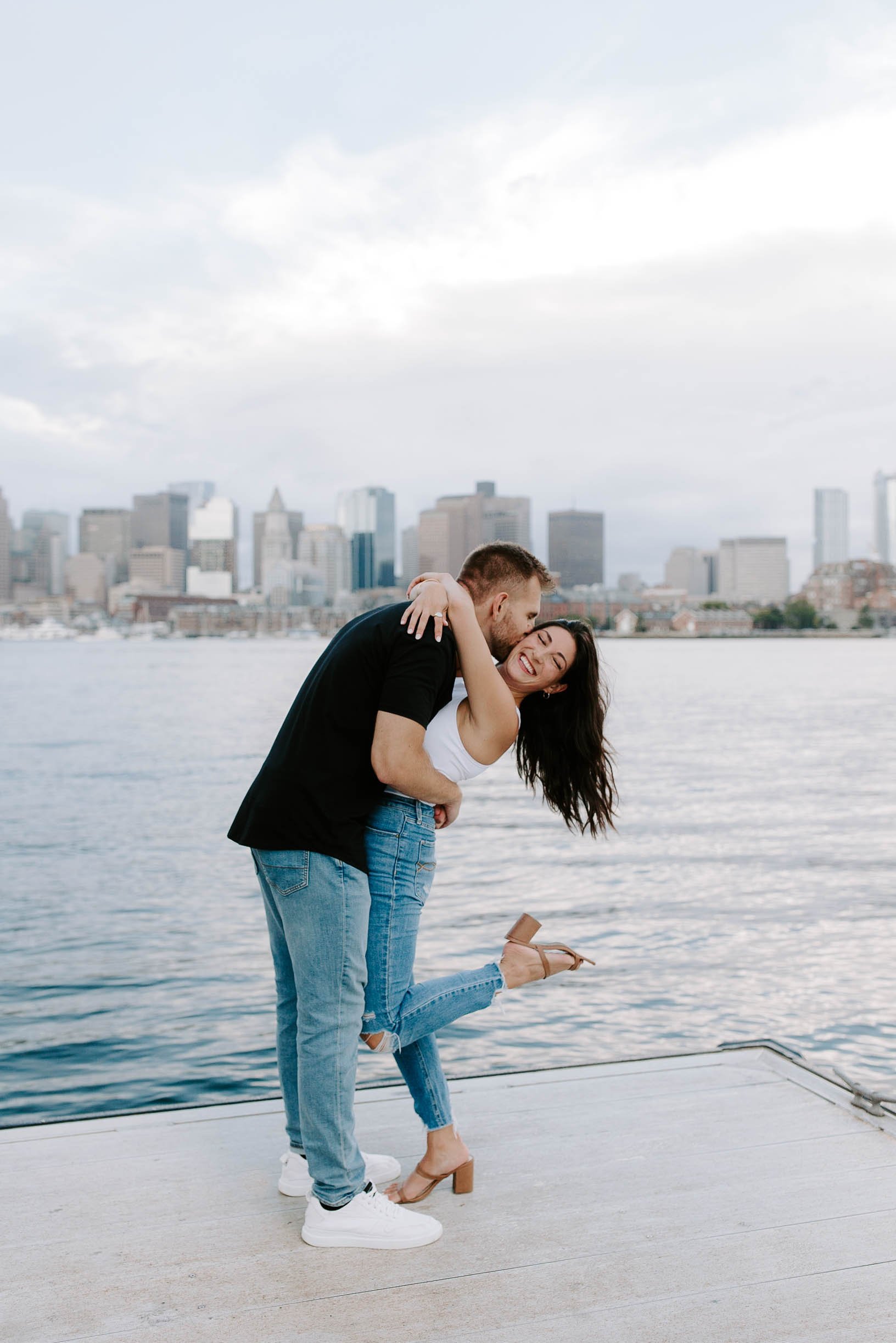  Engaged couple smiling and laughing holding one another at ocean front in front of skyline view  for their boston Seaport engagement session 