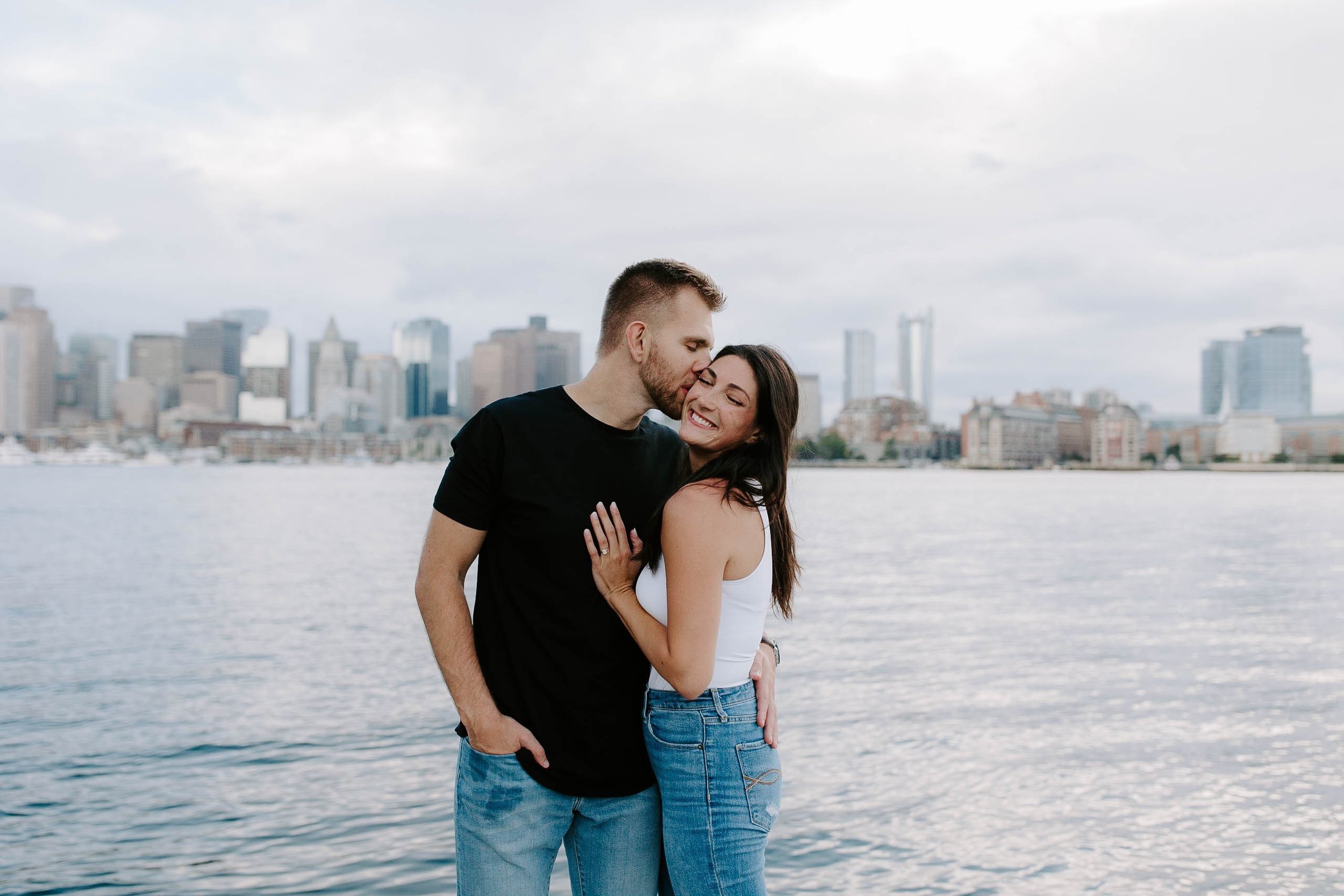 Boyfriend kissing girlfriends forehead while standing on the waterfront with the skyline view in the background