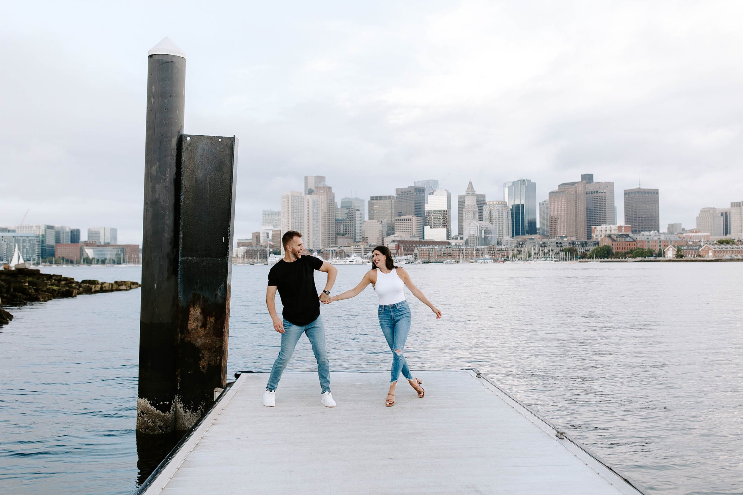  Engaged couple smiling holding hands at Seaport with the skyline view behind them fo their Boston Seaport engagement session 