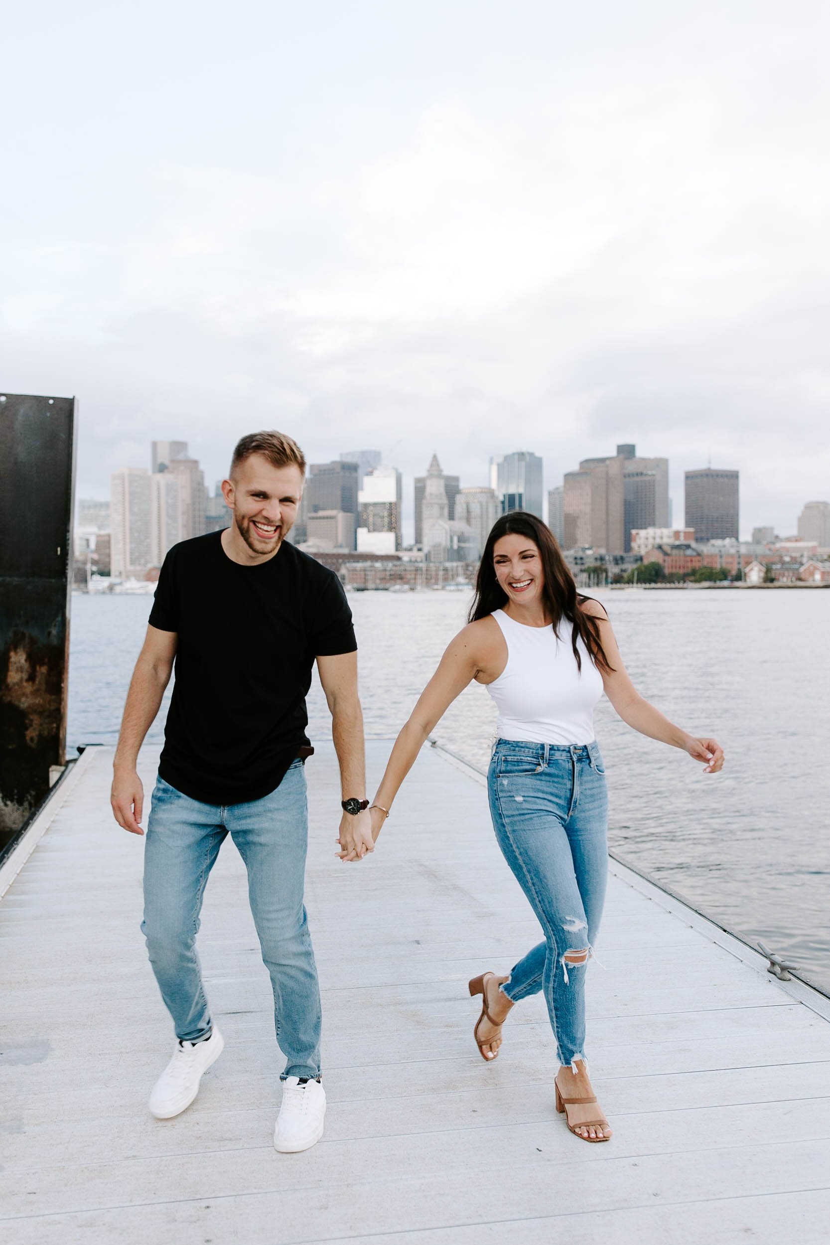  Engaged couple walking and laughing in front of  skyline view  for their boston Seaport engagement session 