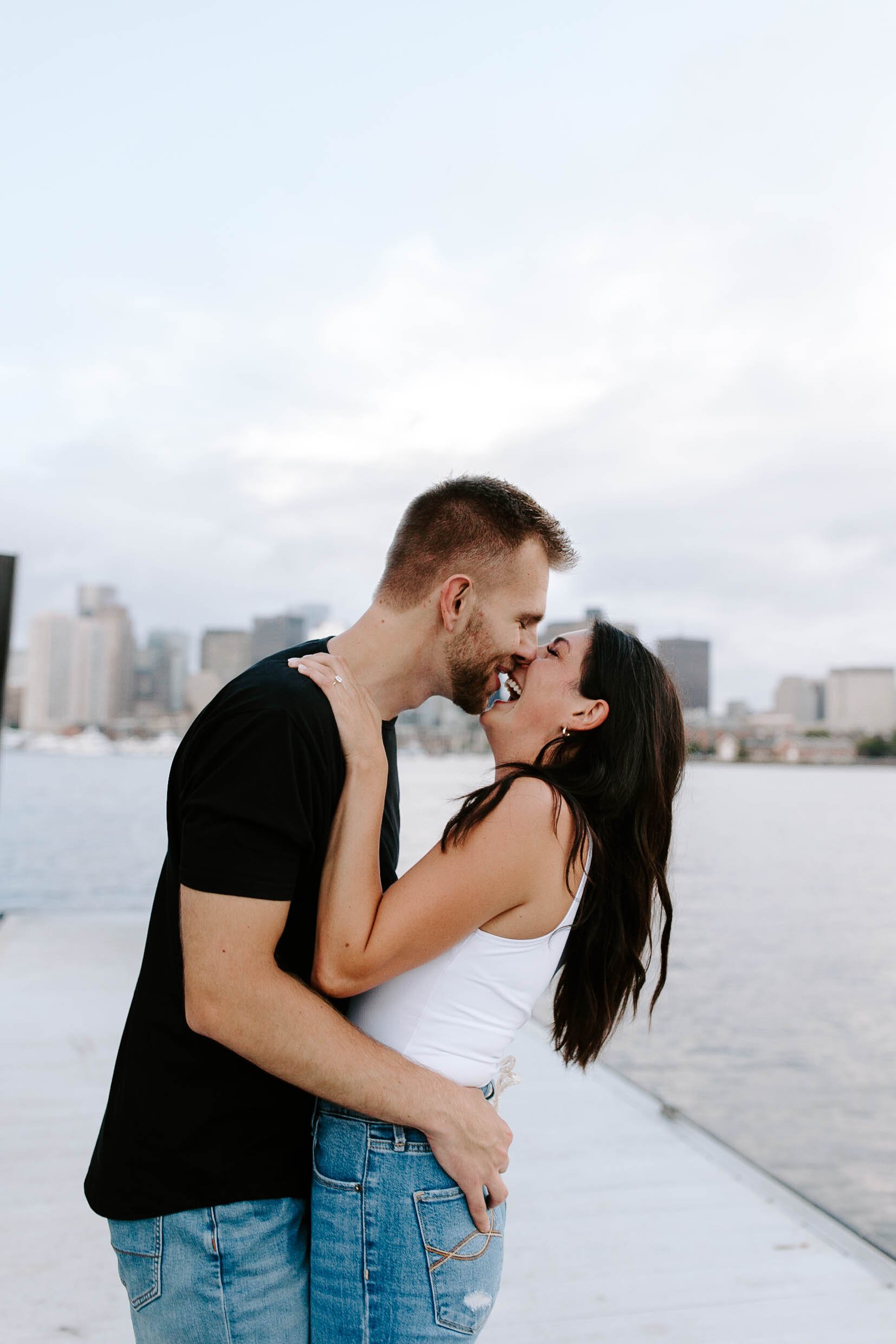  Engaged couple smiling and laughing holding one another at ocean front in front of skyline view  for their Boston Seaport engagement session 