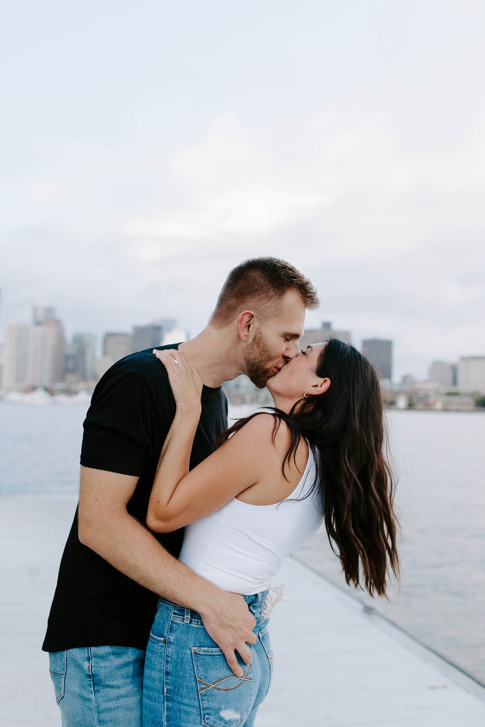  Engaged couple kissing and holding one another at ocean front in front of skyline view  for their boston Seaport engagement session 