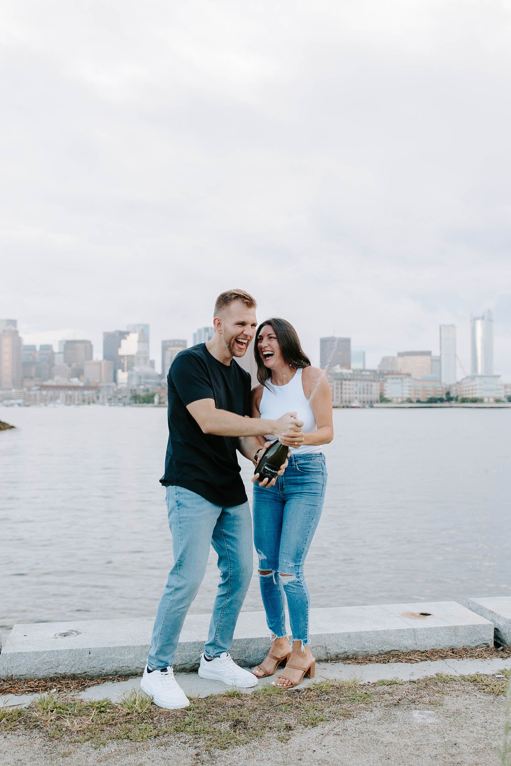  Engaged couple popping a bottle of champagne at ocean front in front of skyline view  for their boston Seaport engagement session 