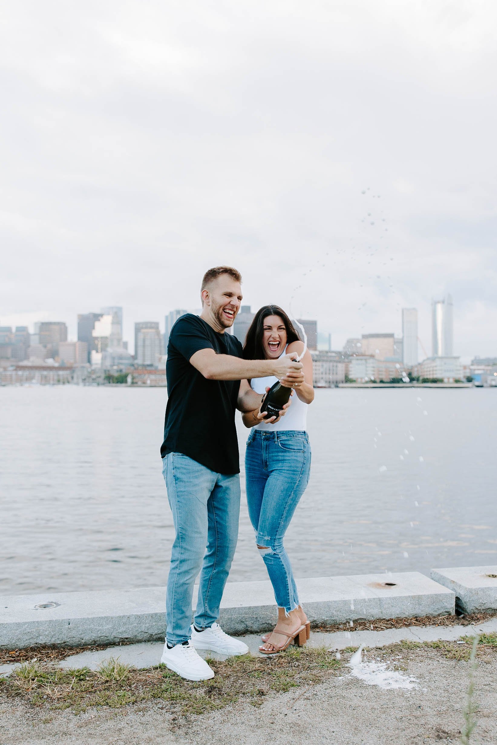  Engaged couple popping champagne at ocean front in front of skyline view  for their boston Seaport engagement session 
