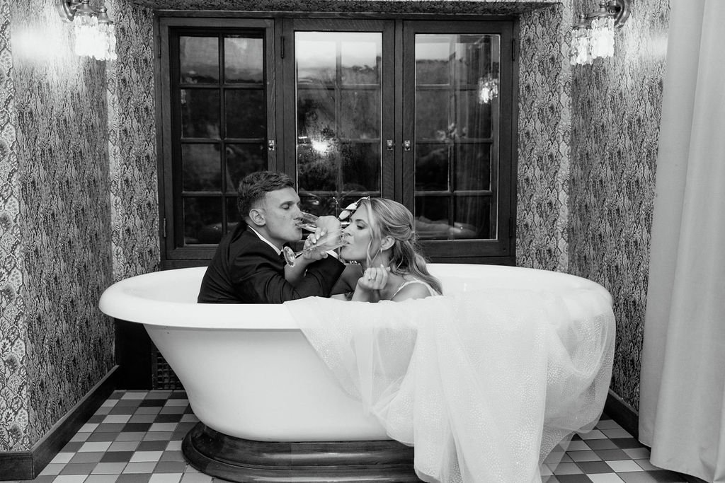 Bride and groom sitting in vintage bathtub drinking champagne