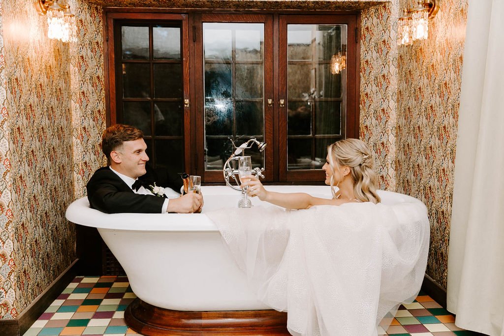 Bride and groom sitting in vintage bathtub smiling at one another holding champagne glasses
