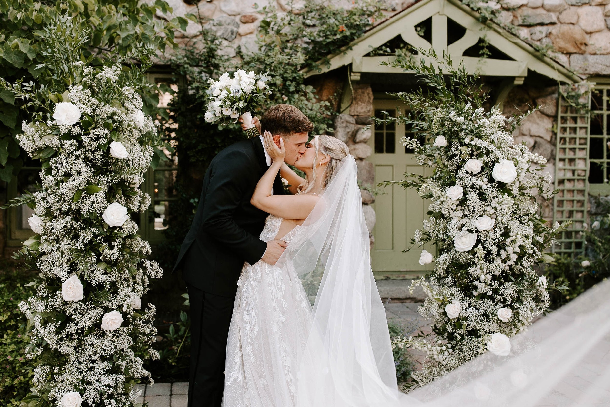Bride and groom kissing after wedding ceremony standing in front of white and green floral arrangements