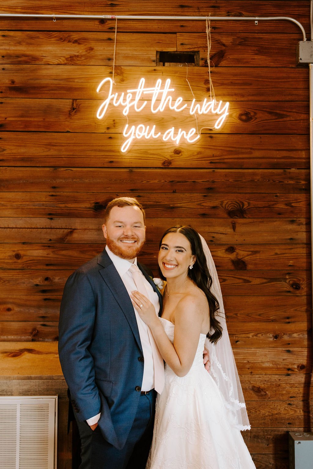 Bride and groom smiling in front of sign that read "Just the way you are"