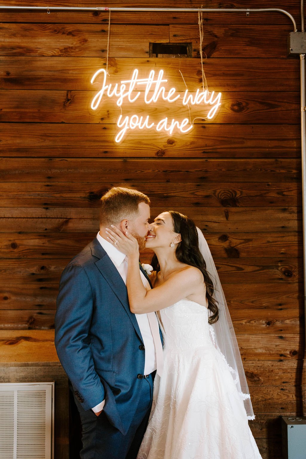 Bride and groom smiling at eachother in front of sign that read "Just the way you are"