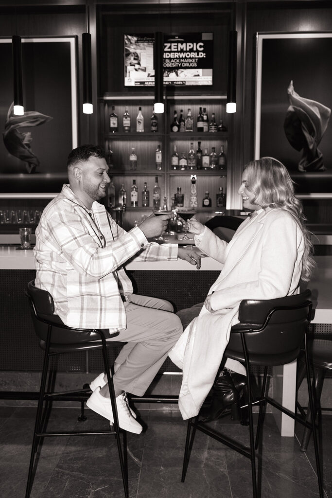 Couple clinking glasses with espresso martinis at a rooftop table during their engagement shoot.