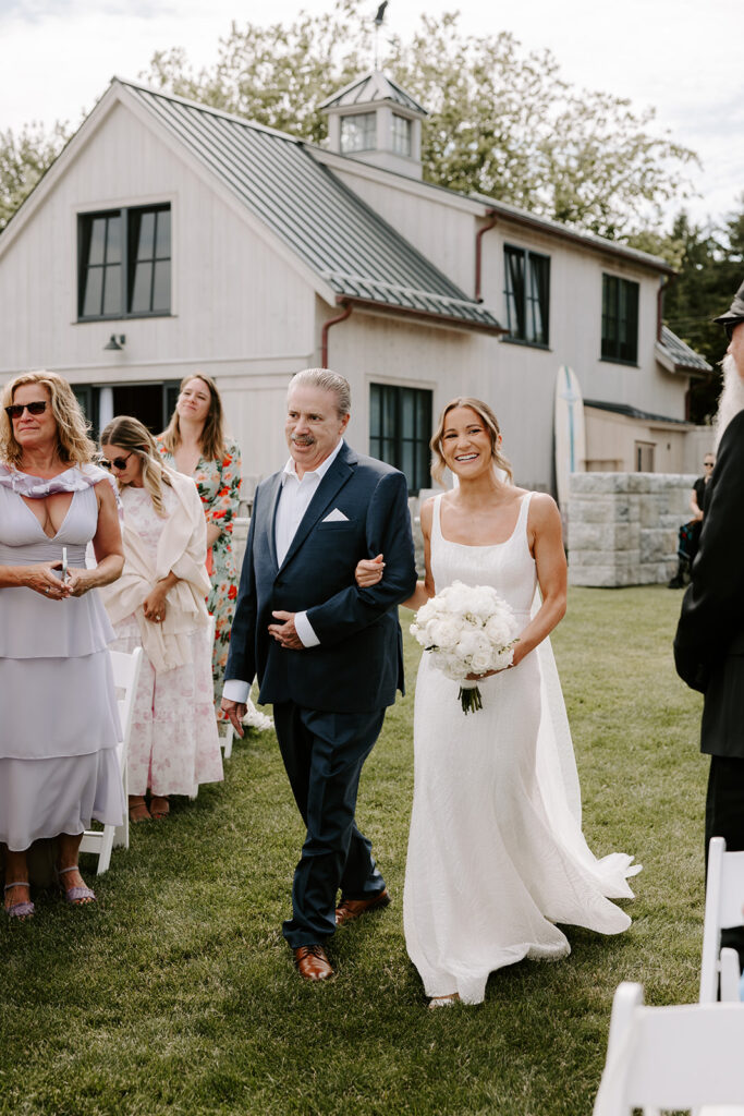 Bride walking down the grassy aisle, holding a bouquet of white roses and greenery