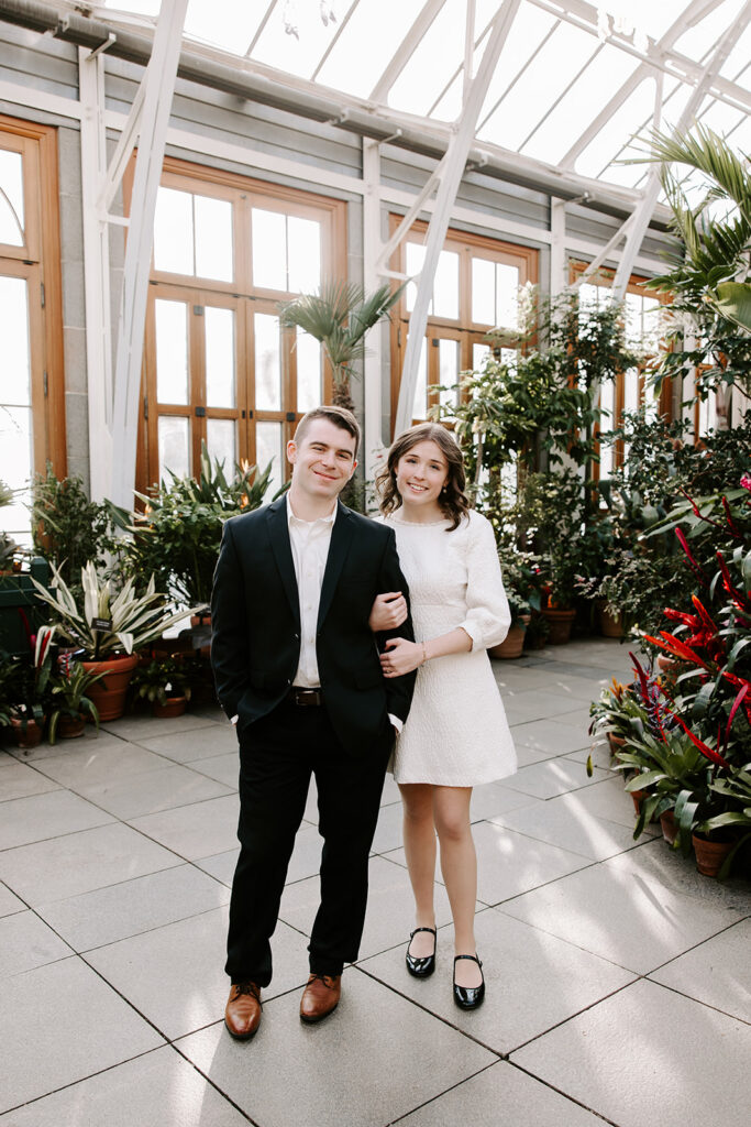 Groom holding fiancée in front of large greenhouse windows with vibrant plants