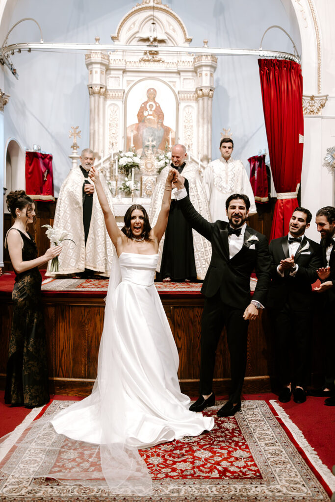 Bride and groom standing beneath the stained glass windows of St. James Armenian Church during their wedding ceremony