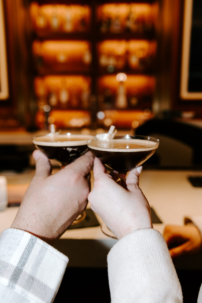 Couple clinking glasses with espresso martinis at a rooftop table during their engagement shoot.
