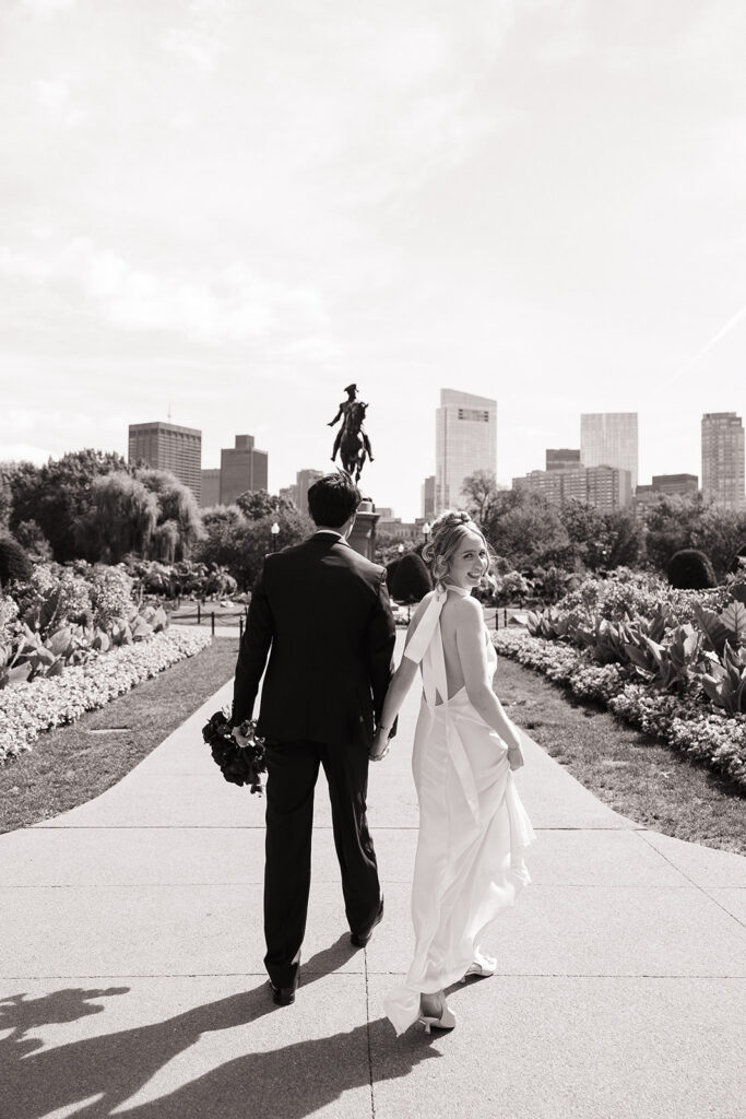 Bride and groom walking toward the George Washington statue in the Boston Public Garden with the Boston skyline behind them