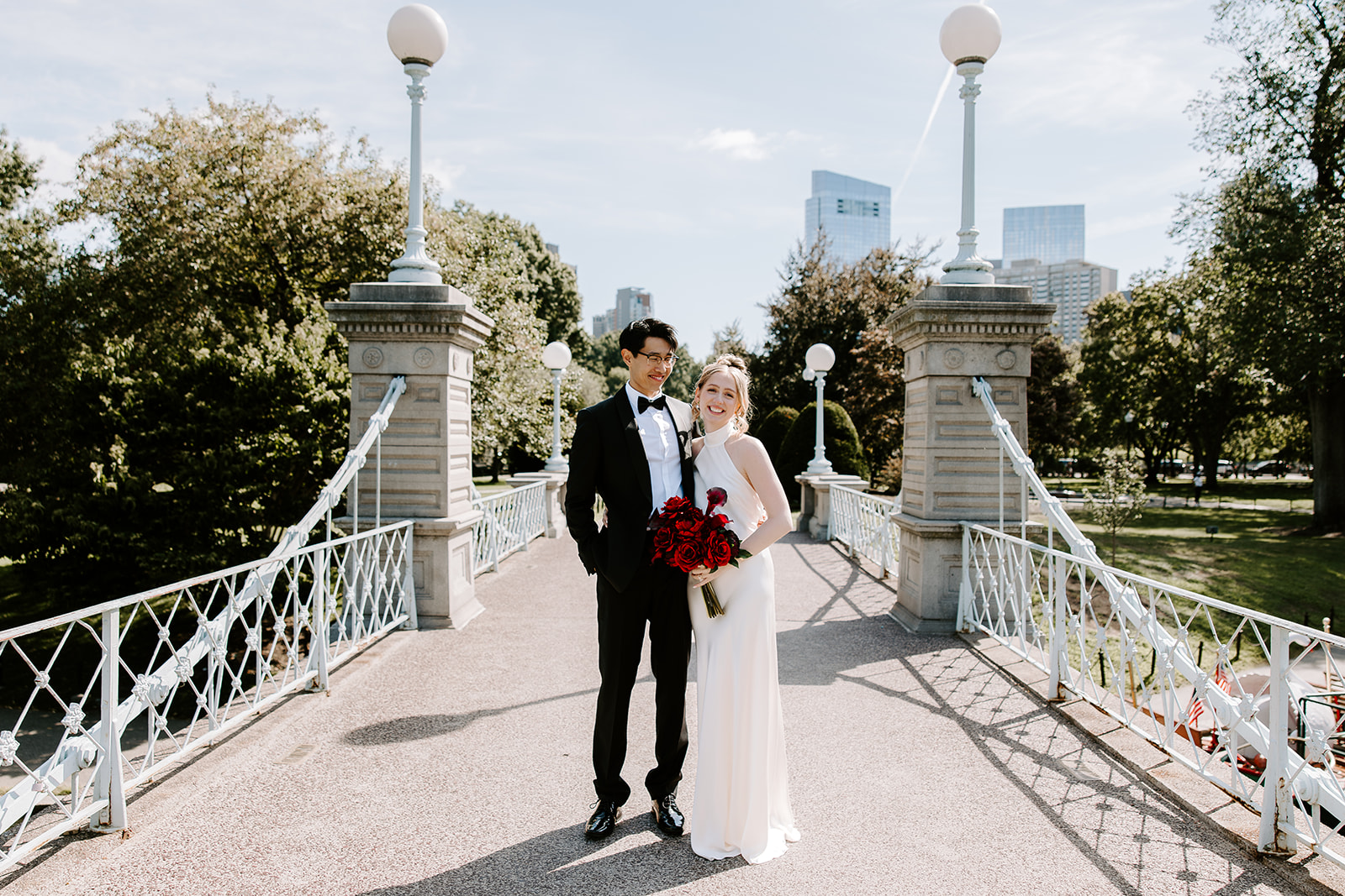 Bride and groom on the Boston Public Garden bridge holding red rose bouquet with city skyline behind them