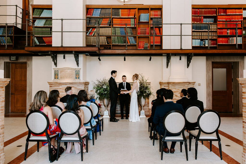 Intimate elopement ceremony in the Boston Public Library with family seated in round-backed chairs and colorful bookshelves behind the couple
