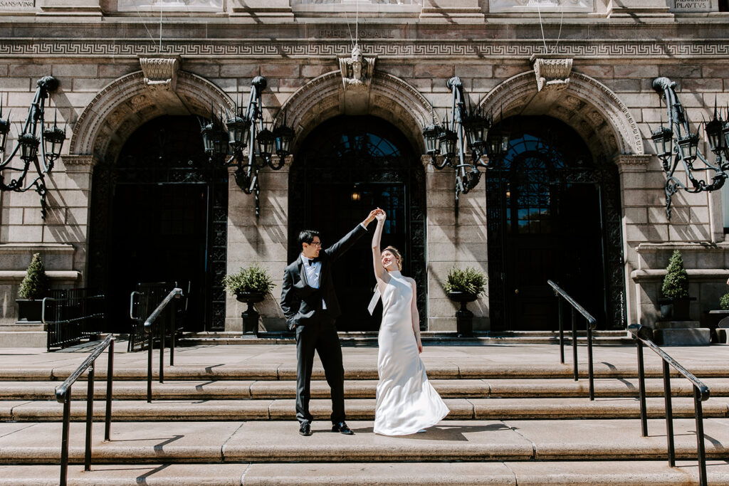 Bride and groom dancing and spinning on the front steps of the Boston Public Library in Copley Square after their intimate elopement ceremony, the iconic stone facade and arched entrances behind them