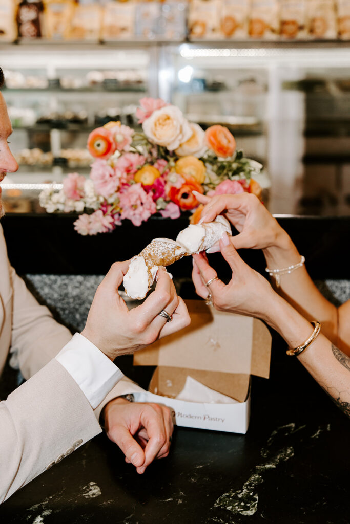 Close up of bride and groom sharing a cannoli from Modern Pastry in Boston's North End with wedding rings and bridal bouquet visible, photographed by Genevieve Photography