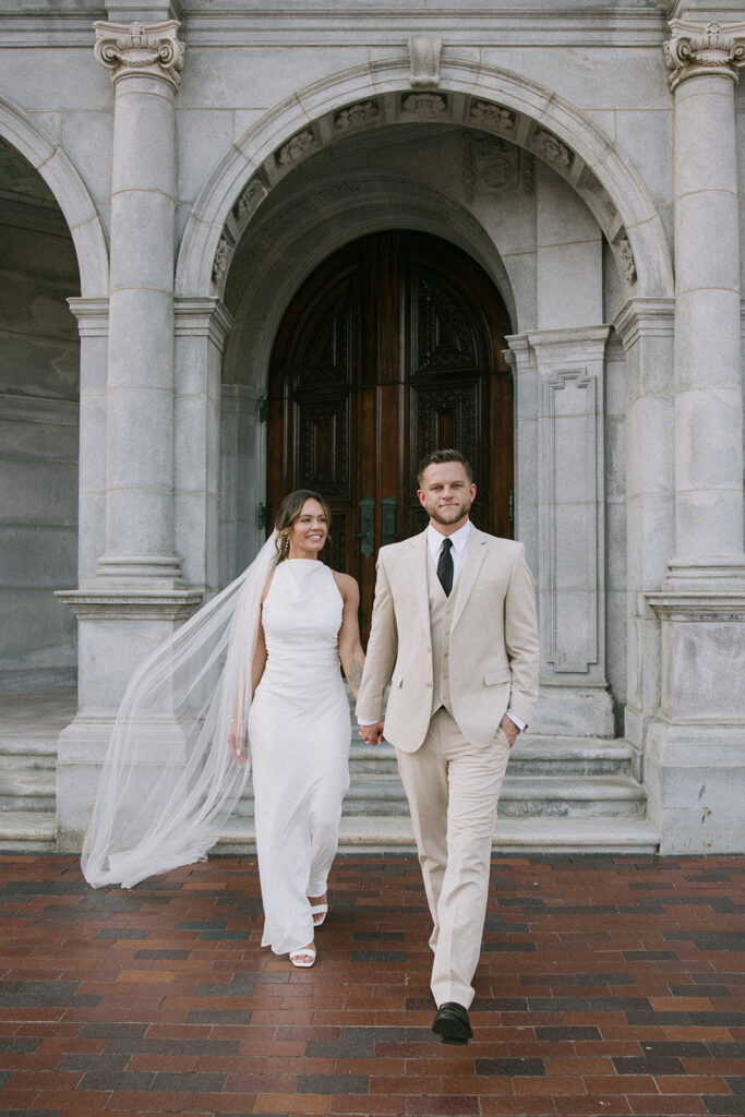 Bride and groom walking hand in hand under the ornate stone arch entrance of the First Church of Christ Scientist at the Christian Science Center in Boston, Boston City Hall wedding portrait photographed by Boston wedding photographer Genevieve Photography