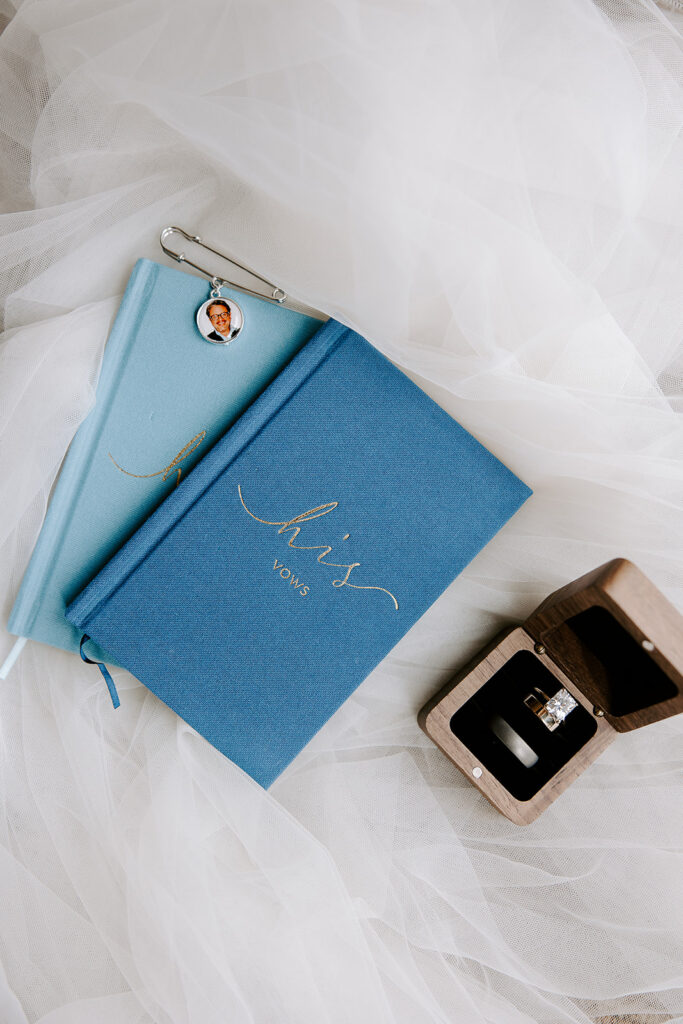 Wedding detail flat lay with blue vow book, memorial charm bouquet pin, and wooden ring box with diamond engagement ring and wedding band, Boston City Hall wedding photographed by Genevieve Photography