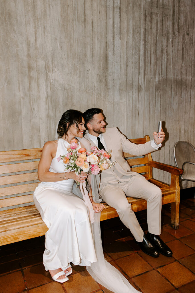 Bride and groom taking a selfie on a bench inside Boston City Hall after their intimate wedding ceremony, bride holding colorful bridal bouquet, photographed by Boston wedding photographer Genevieve Photography