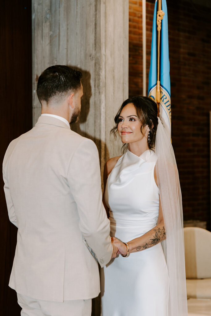 Bride gazing at groom during wedding vows at Boston City Hall Room 222 with the Massachusetts state flag behind them, intimate Boston City Hall wedding photographed by Genevieve Photography