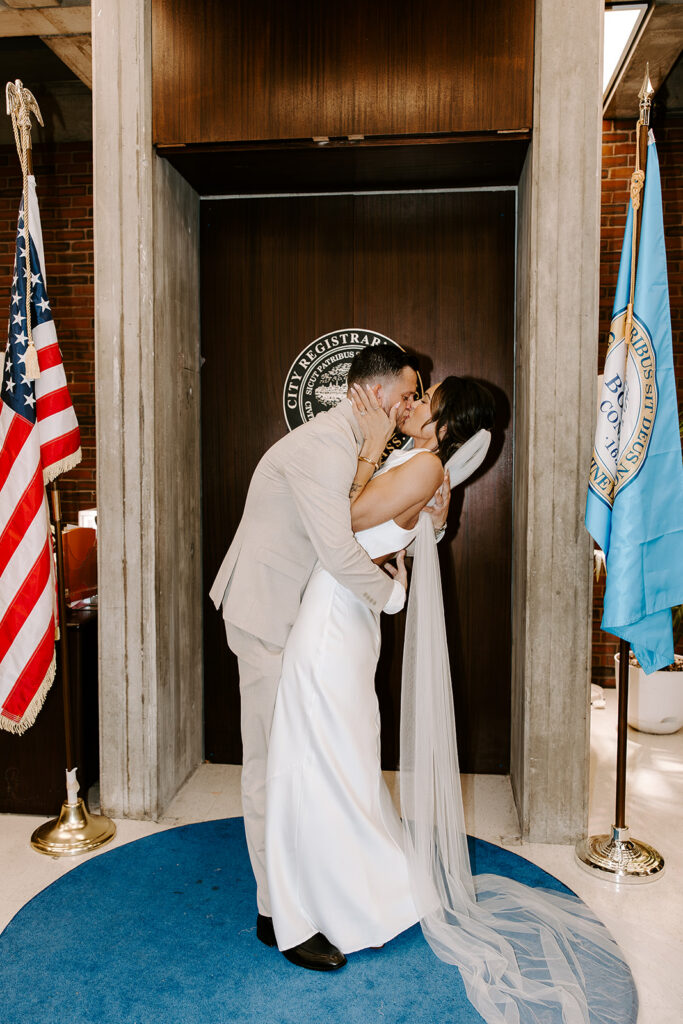 Bride and groom first kiss at Boston City Hall in the City Registrar's office with the Boston City seal behind them and cathedral veil spread on the blue carpet, photographed by Genevieve Photography