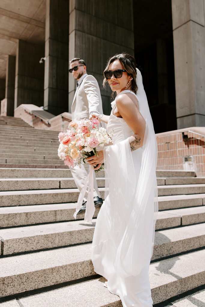 Bride in white satin halter gown with cathedral veil looking back on the steps of Boston City Hall, Boston wedding photographer Genevieve Photography