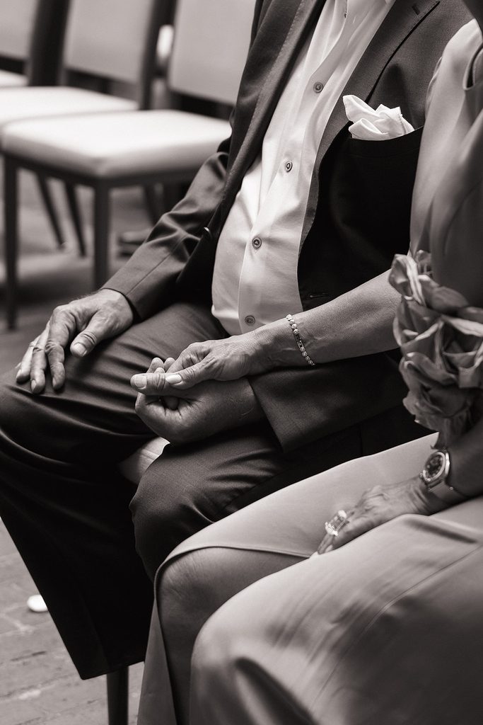 Black and white photo of two wedding guests holding hands as they watch the ceremony in the courtyard at the Four Seasons Hotel Boston, intimate Boston wedding photographed by Boston elopement photographer Genevieve Photography