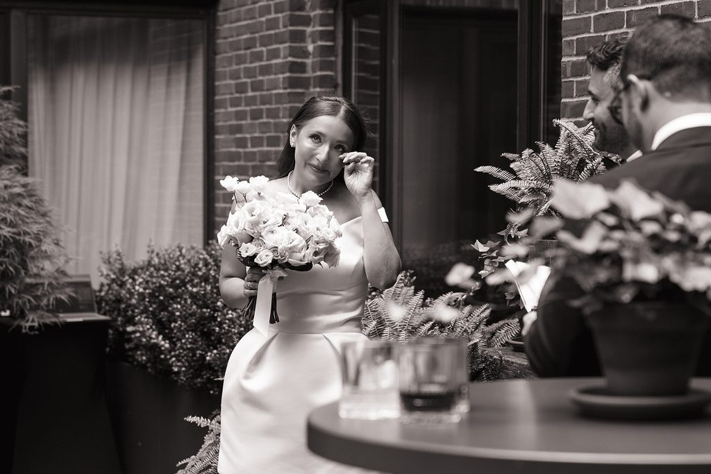 Black and white photo of a bride wiping away a happy tear while holding a bouquet of white roses during her courtyard wedding ceremony at the Four Seasons Hotel Boston, photographed by Boston elopement photographer Genevieve Photography