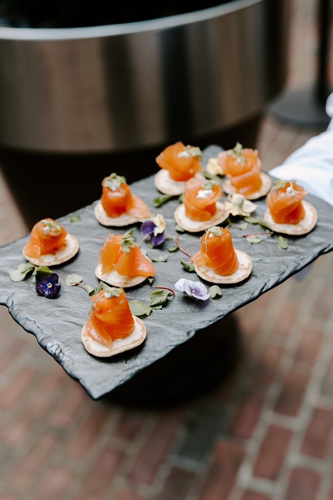 Smoked salmon bites on mini pancakes with fresh herbs and edible flowers, served on a slate tray during the cocktail hour of an intimate wedding at the Four Seasons Hotel Boston, photographed by Boston elopement photographer Genevieve Photography