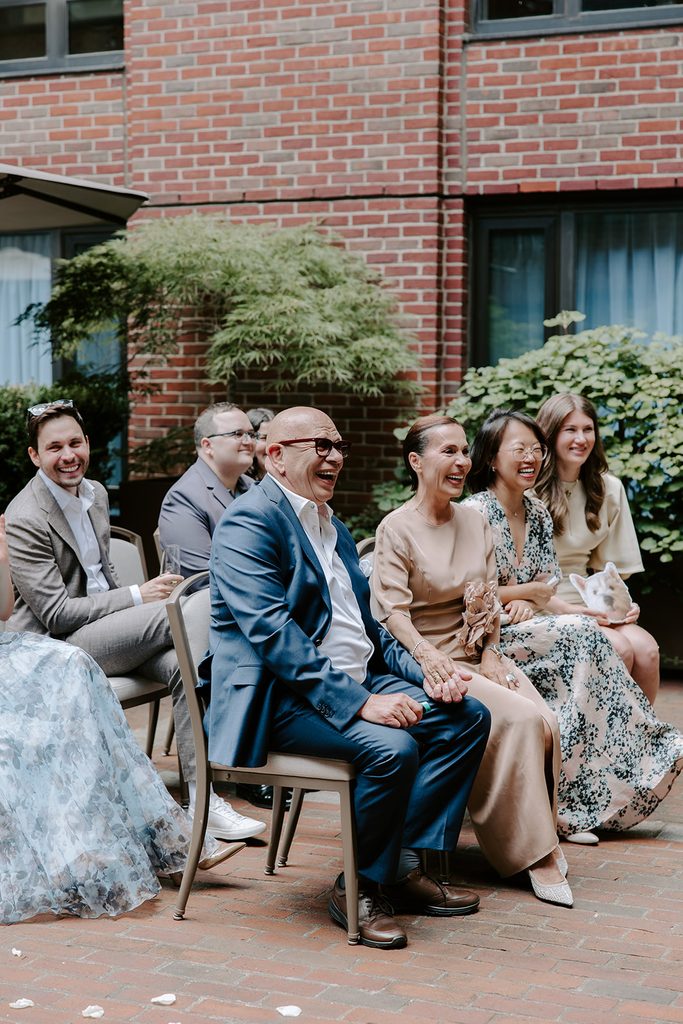 Wedding guests laughing together during a courtyard ceremony at the Four Seasons Hotel Boston, surrounded by red brick and greenery, intimate Boston wedding photographed by Boston elopement photographer Genevieve Photography