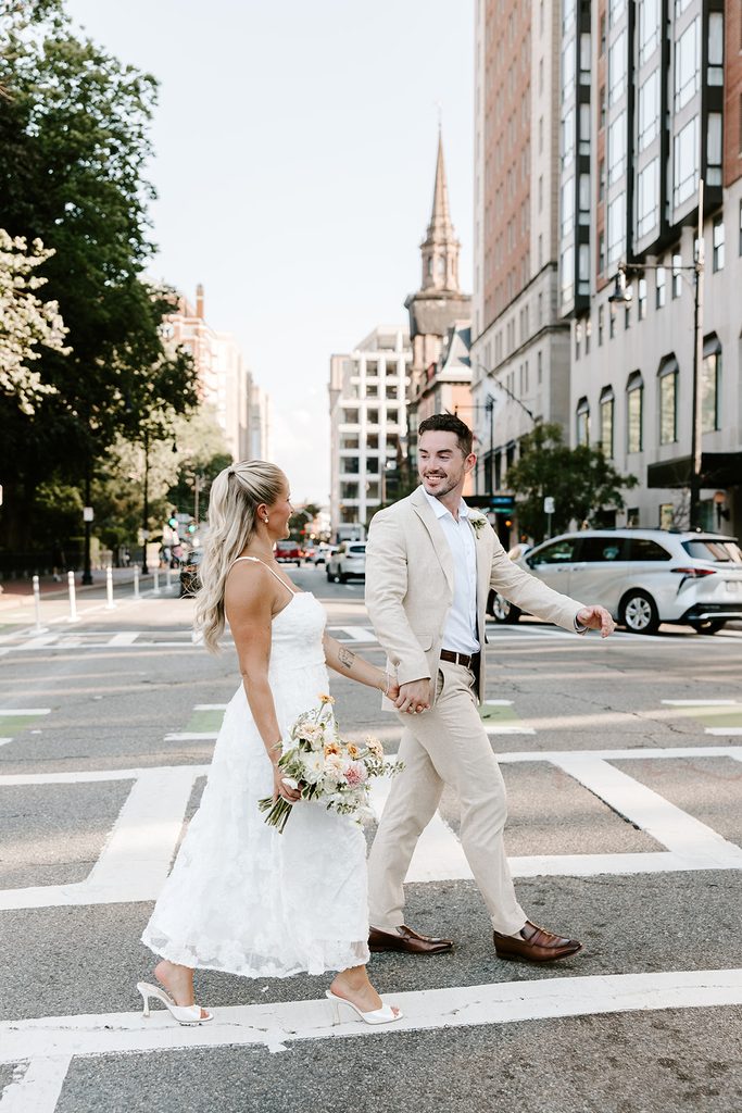 Groom smiling back at his bride as he leads her by the hand across a crosswalk in Boston, a church steeple and city buildings behind them, after their Boston Public Garden elopement, photographed by Boston elopement photographer Genevieve Photography