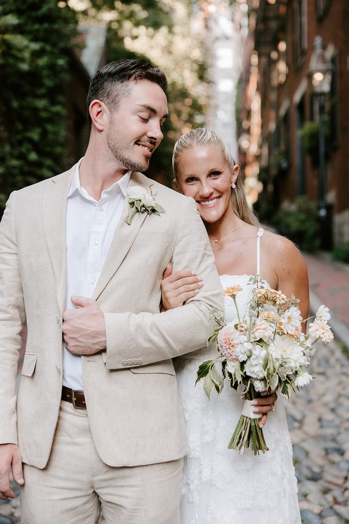 Bride smiling and resting her head on her groom's shoulder while holding a peach and white bouquet on the cobblestones of Acorn Street in Beacon Hill, Boston elopement photographed by Boston elopement photographer Genevieve Photography