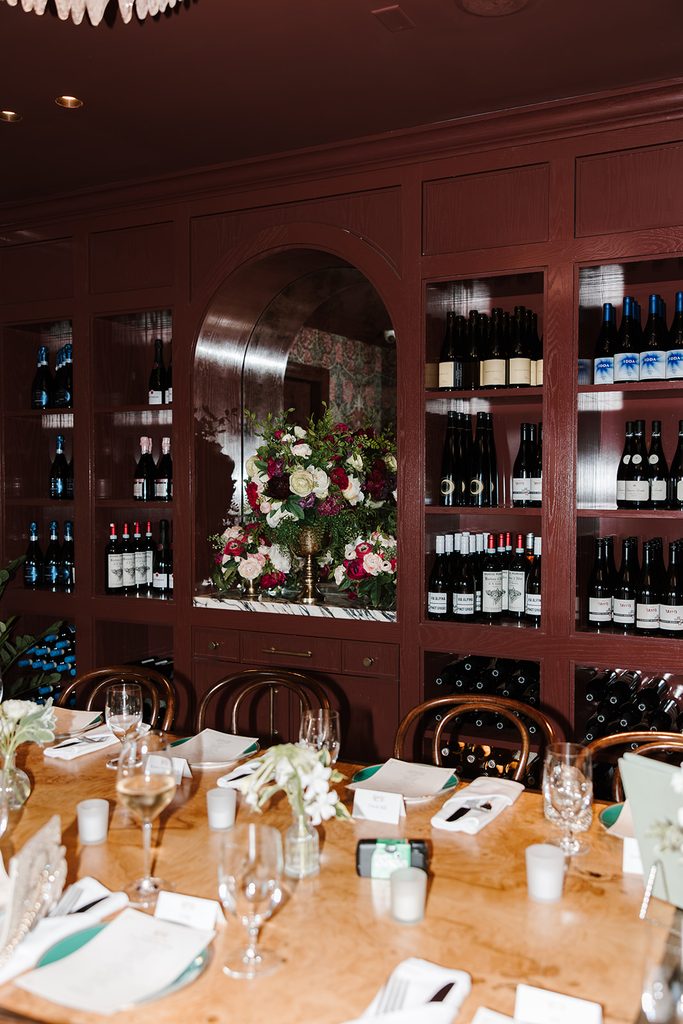 Elopement dinner table set with candles and white flowers in the private dining room at Contessa Boston, with burgundy wine shelves and a large red and white floral arrangement in an arched niche, photographed by Boston elopement photographer Genevieve Photography