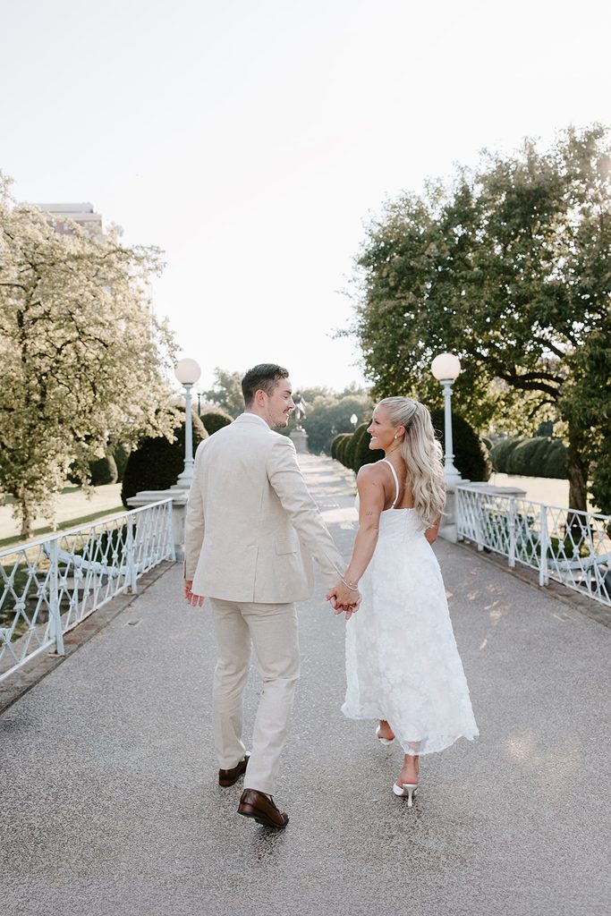 Bride and groom holding hands and smiling at each other as they walk across the Boston Public Garden footbridge, with the George Washington statue ahead of them. Boston Public Garden elopement photographed by Boston elopement photographer Genevieve Photography