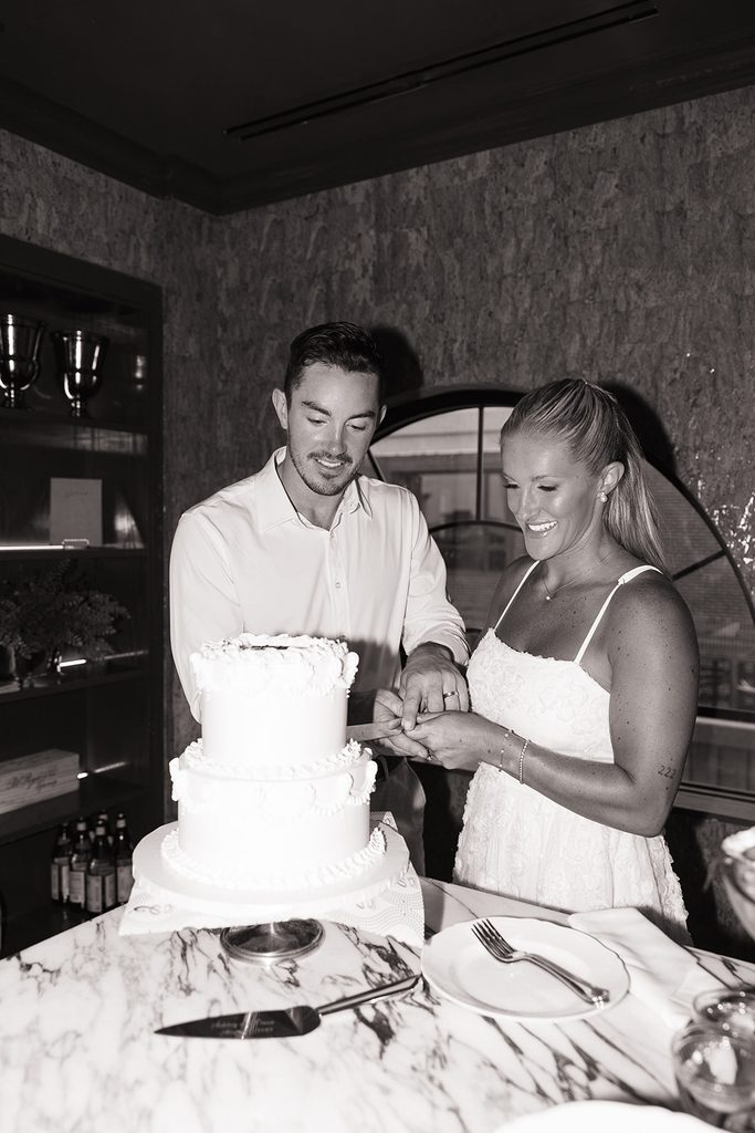 Black and white photo of a bride and groom smiling as they cut their tiered white wedding cake together at their elopement dinner at Contessa Boston, photographed by Boston elopement photographer Genevieve Photography