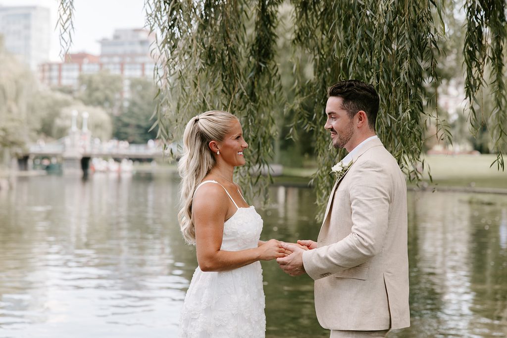 Bride and groom holding hands and smiling at each other under a weeping willow by the Boston Public Garden lagoon, with the footbridge and swan boats behind them. Intimate Boston Public Garden elopement photographed by Boston elopement photographer Genevieve Photography