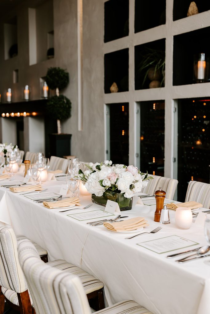 Long reception table set with white linens, gold napkins, glowing candles, and low white rose centerpieces beside a wall of wine at Mistral in Boston's South End, restaurant wedding photographed by Boston elopement photographer Genevieve Photography