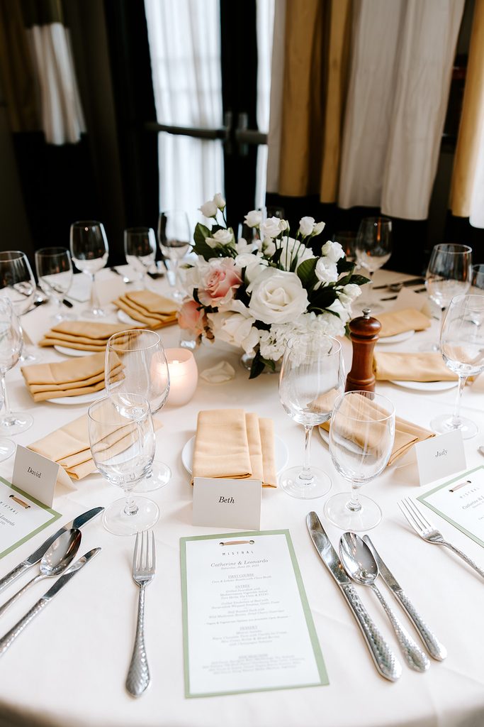 Round reception table at Mistral in Boston with a custom wedding menu for Catherine and Leonardo, name cards, gold napkins, and a white and blush rose centerpiece, restaurant wedding photographed by Boston elopement photographer Genevieve Photography