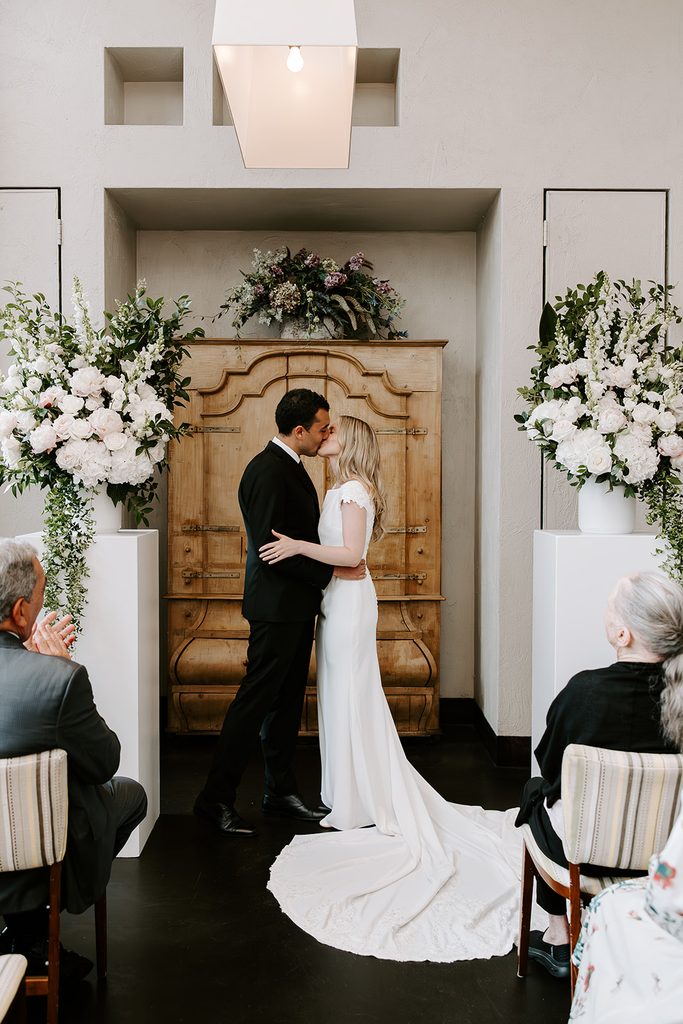 Bride and groom sharing their first kiss in front of an antique wooden armoire with tall white floral arrangements on each side, her long train spread on the floor, at their restaurant wedding ceremony at Mistral in Boston, photographed by Boston elopement photographer Genevieve Photography