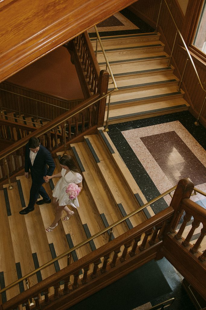 Overhead view of a bride holding pink peonies and her groom walking hand in hand down the grand wooden staircase at Cambridge City Hall, with brass railings and a patterned stone floor below. Cambridge City Hall wedding photographed by Boston elopement photographer Genevieve Photography