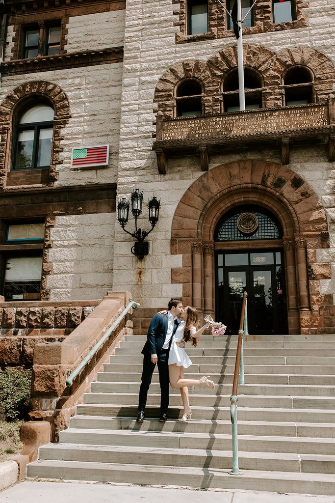 Bride raising her pink peony bouquet and kicking up her heel as she kisses her groom on the front steps of Cambridge City Hall, with the stone arched entrance and iron lantern behind them. Cambridge City Hall wedding photographed by Boston elopement photographer Genevieve Photography
