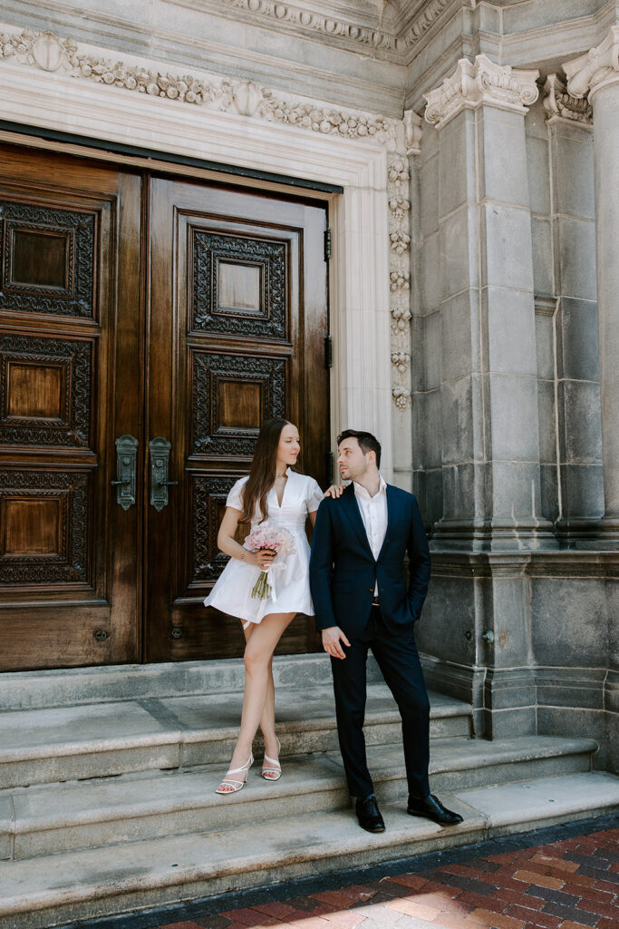 Bride in a short white dress with pink peonies resting her hand on her groom's shoulder in front of tall carved wooden doors and stone columns near the Christian Science Plaza in Boston, after their Cambridge City Hall wedding, photographed by Boston elopement photographer Genevieve Photography
